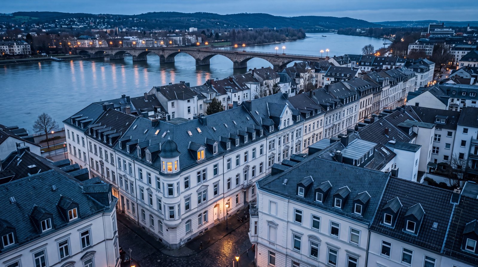 Luftbild einer Stadt am Rhein in der blauen Stunde: Fluss, Steinbogenbrücke, Gründerzeitviertel, ein Eckhaus mit Turm und einem einzigen erleuchteten Fenster
