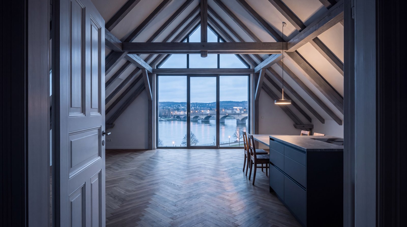 Wohnküche unter dem Dach mit offenem Dachstuhl, raumhohem Giebelfenster und Blick auf Fluss und Brücke